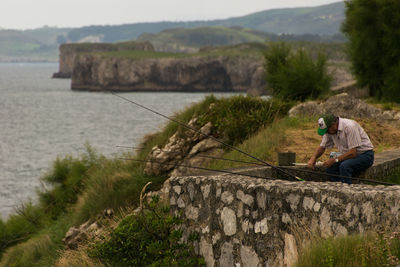 Man sitting on rock by sea against mountains