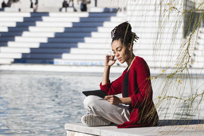 Young woman sitting cross-legged using digital tablet while at edge of promenade over river on sunny day