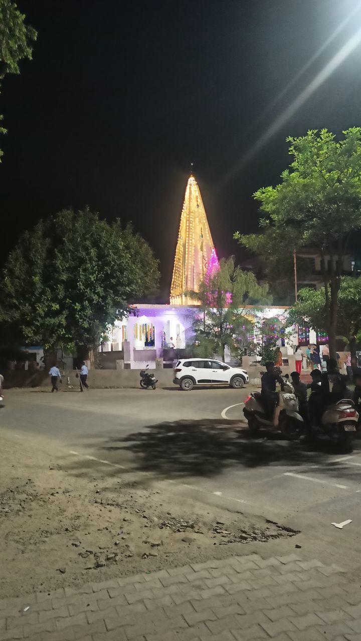 tree, architecture, night, plant, city, nature, street, illuminated, sky, group of people, light, built structure, transportation, mode of transportation, celebration, crowd, car, outdoors, lighting, large group of people, building exterior, event, vehicle, motor vehicle