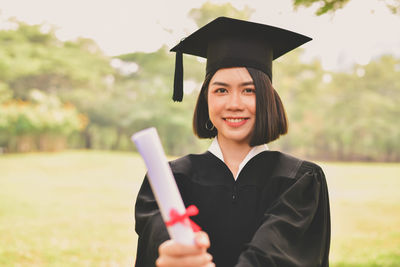 Portrait of young woman in graduation gown holding certificate while standing at park