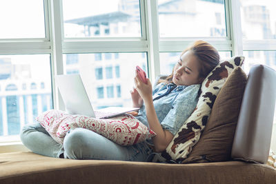 Young woman using mobile phone while sitting on window