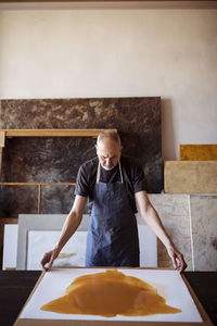 Confident senior artist holding painting at table in workshop