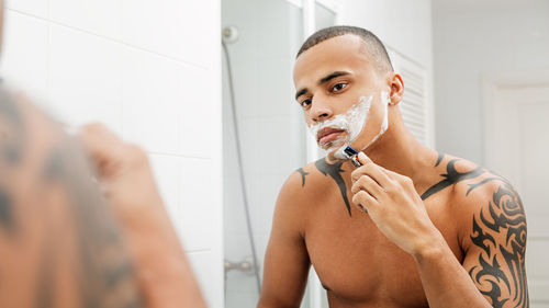 Tattooed shirtless man shaving while looking in mirror