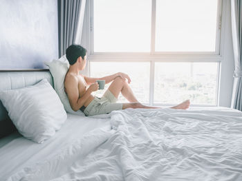 Asian young man with tattoo holding coffee cup on the bed looking out window in morning with sunrise