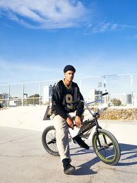 Young ethnic hipster male athlete in cool wear sitting on bmx bike while looking away in town on sunny day