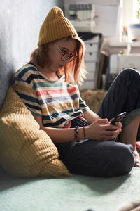 Side view of female millennial in trendy wear sitting on sofa at home and chatting with friends on social media via mobile phone