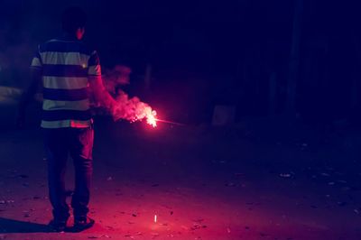 Rear view of man holding burning sparkler on field at night