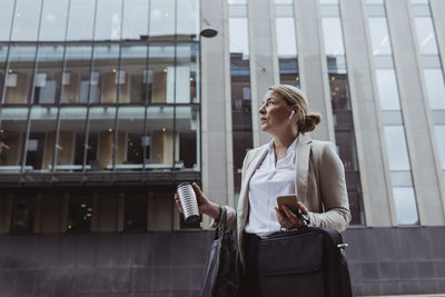Female entrepreneur with bag and coffee cup looking away while standing against building in city