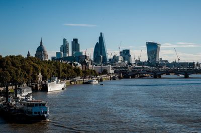 Boats in river by cityscape against blue sky