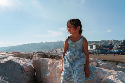 Side view of young girl sitting on rock against sky