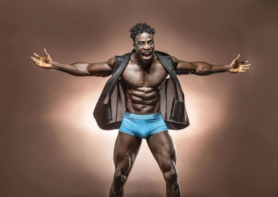 Portrait of shirtless athlete with arms outstretched standing against brown background