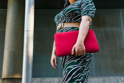 Side view of anonymous cheerful young female in stylish striped outfit with red laptop case in hands smiling while standing against contemporary city building