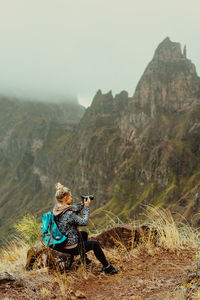 Rear view of woman photographing while sitting against mountain