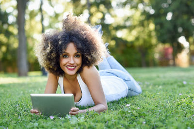 Portrait of smiling young woman with digital tablet lying on grassy field at public park