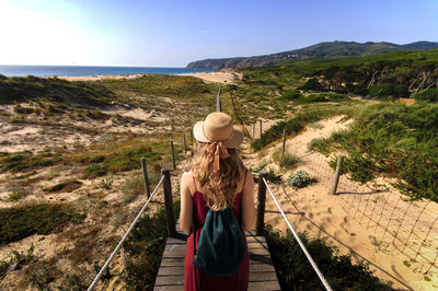 Rear view of young woman with backpack standing on boardwalk at beach against clear sky during sunny day