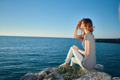 Woman sitting on rock by sea against clear sky