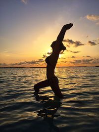 Silhouette woman with arms outstretched exercising at beach against sky during sunset