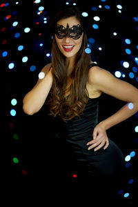 Portrait of beautiful young woman wearing mask standing against illuminated lights