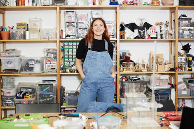 Portrait of smiling redhead engineer standing with hands in pockets against shelf at workshop