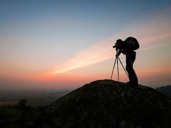 Man photographing on rock against sky during sunset