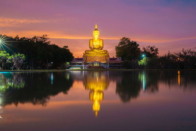 A large buddha image is illuminated by the lake against the sky during sunset.