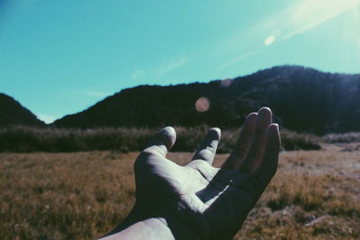 Cropped hand over grassy field against blue sky during sunny day