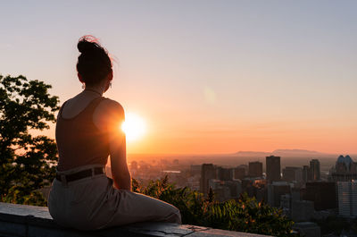 Silhouette woman sitting by cityscape against sky during sunset