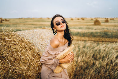 Smiling woman in sunglasses with bare shoulders on a background of wheat field and bales of hay.