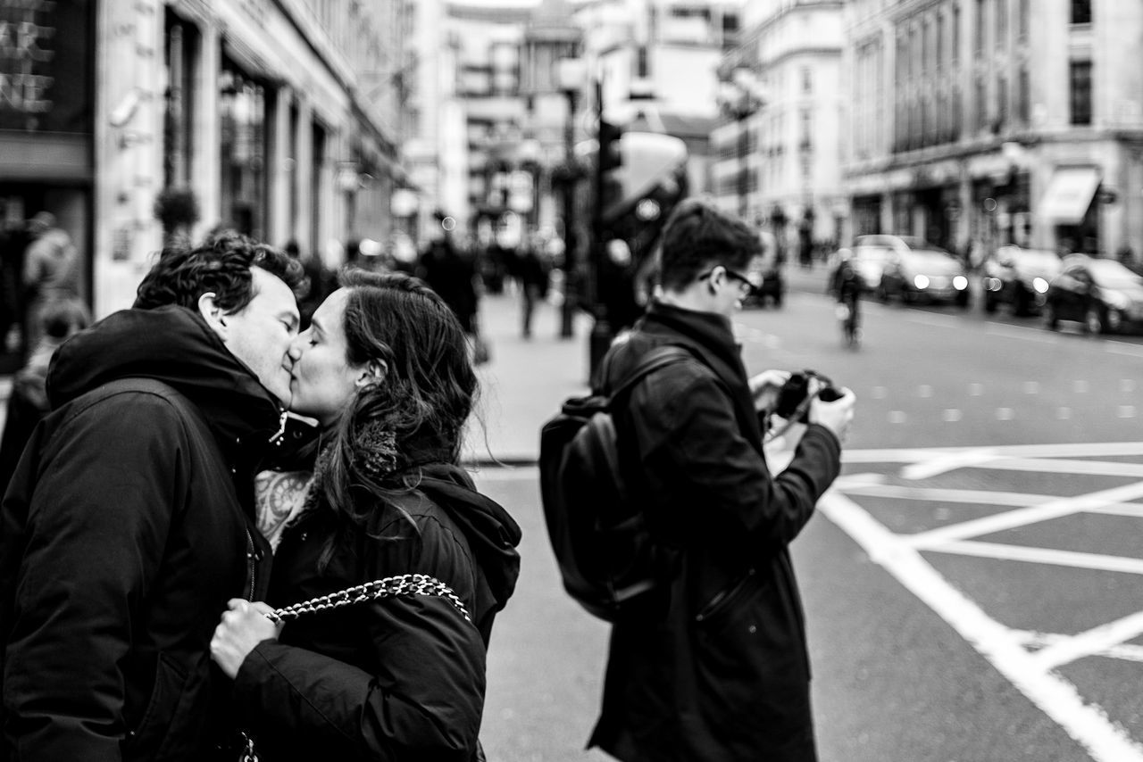 city, real people, two people, street, lifestyles, people, women, focus on foreground, architecture, men, togetherness, building exterior, adult, technology, photography themes, built structure, incidental people, waist up, day, photographing, outdoors, warm clothing, couple - relationship