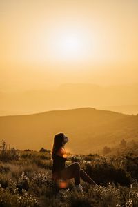 Side view of man sitting on landscape against sky at sunset