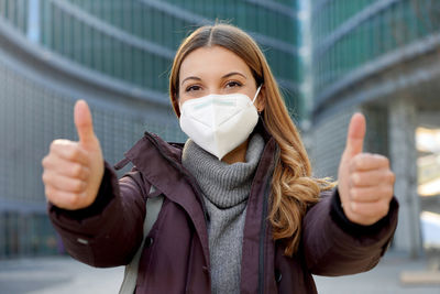 Girl wearing protective face mask showing thumbs up in modern city street and looking at camera