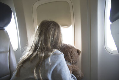 Rear view of girl with teddy bear looking through window while traveling in airplane