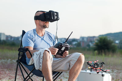 Mid adult man using virtual reality simulator while sitting on chair against sky