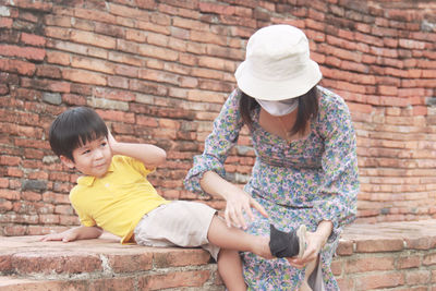 Full length of mother and girl sitting against brick wall