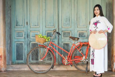 Portrait of smiling young woman with hat standing by bicycle against closed door