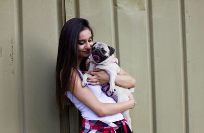 Smiling young woman holding pug while standing against corrugated iron