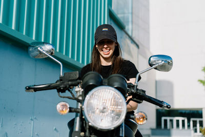 Positive female motorcyclist sitting on motorbike parked in city street on sunny day and looking at camera