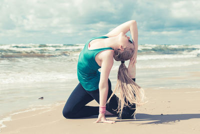 Side view of young woman doing yoga at beach against cloudy sky
