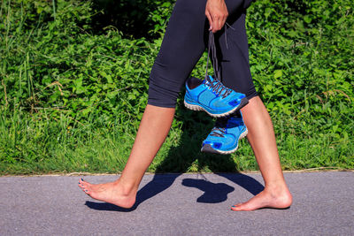 Low section of woman holding sports shoes while walking on road during sunny day