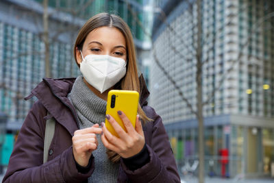 Woman with medical face mask checking notifications on smartphone on urban background. copy space.