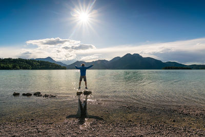 Man with arms outstretched jumping on rock in lake against sky