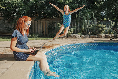 Woman using laptop while girl jumping in swimming pool