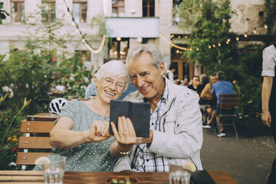 Smiling woman taking selfie with senior man while sitting at restaurant in city
