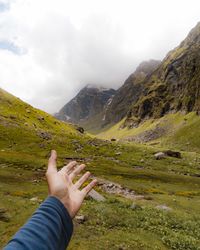 Cropped hand gesturing towards mountain against cloudy sky