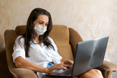 Cute woman at home in chair with laptop wearing protective mask on her face