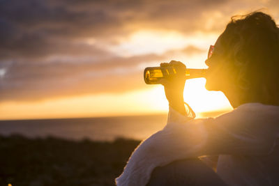 Side view of mature woman drinking while sitting against cloudy sky during sunset