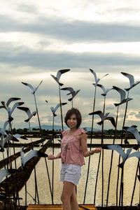 Portrait of mid adult woman standing by sculptures by lake against cloudy sky during sunset