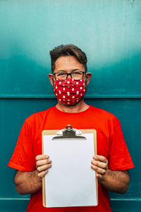 Portrait of man wearing mask holding clipboard against blue wall