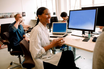 Smiling businesswoman having drink looking away while sitting with colleague at office desk