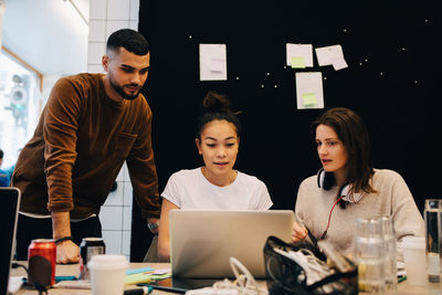 Confident young businesswoman discussing with multi-ethnic colleagues over laptop against bulletin board at office